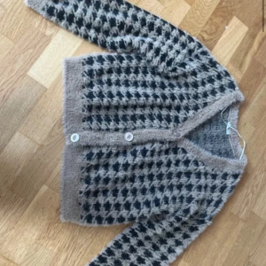 Rutig kofta från Stories i beige och svart - Supermjuk och fluffig kofta från Stories i storlek S. Koftan har ett rutigt mönster i beige och svart, v-ringning och knappar framtill. 