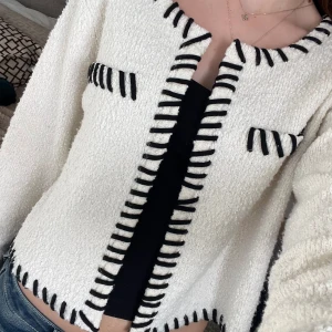 Vit kofta med svarta detaljer - Säljer en vit kofta från H&M med grovstickad känsla och svarta dekorativa sömmar längs kanter, fickor och halsringning. Koftan är croppad, har långa ärmar och en boxig passform. Perfekt för att lyfta en enkel outfit.