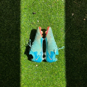 Nike blå fotbollsskor med orange detaljer - Säljer ett par Nike fotbollsskor i blått med orange och röda detaljer. Skorna har snörning, rund tå och mönstrad ovandel. Sulan är blå med orange inslag och dobbar för bra grepp på planen. Perfekta för dig som vill sticka ut på fotbollsplanen.