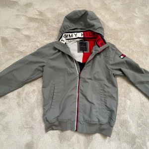 Grå vindjacka från Tommy Hilfiger - Snygg grå vindjacka från Tommy Hilfiger med huva och dragkedja framtill. Jackan har ribbade muddar, två fickor och logotyp på ärmen. Insidan har meshfoder och detaljer i rött, vitt och marinblått. Perfekt för blåsiga dagar.