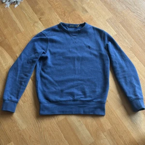 Blå sweatshirt från Polo Ralph Lauren XS - Blå sweatshirt från Polo Ralph Lauren i storlek XS. Tröjan har rund hals, långärmade ärmar och ett diskret broderat logomärke på bröstet. Tillverkad i mjuk bomull med klassisk passform och ribbade muddar vid ärmslut och nederkant.