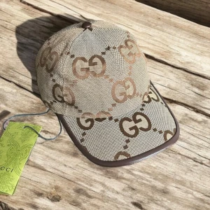 Gucci keps med GG-mönster - Snygg keps från Gucci i beige och brunt med det klassiska GG-logomönstret över hela kepsen. Kepsen har bruna detaljer i skinn runt skärmen och justerbar rem bak. Tillverkad i bomullsmix med exklusiv känsla och tydlig logga.