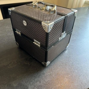 Sminkväska i svart från SOHO - Snygg och rymlig sminkväska från SOHO i svart med silverdetaljer och metallhörn. Väskan har flera utdragbara fack och handtag i metall, perfekt för att organisera smink och borstar. Insidan är mönstrad med SOHO New York-tryck.