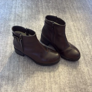 Bruna boots i skinn med dragkedja - Snygga mörkbruna boots i skinn med rund tå och låg klack. Skorna har dragkedja på sidan och ett dekorativt spänne baktill. Perfekta för höst och vinter och passar till många olika outfits.