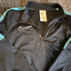 Svart Nike half zip träningströja - Svart långärmad träningströja från Nike med turkosa detaljer längs ärmarna och vit broderad swoosh på bröstet. Tröjan har en praktisk half zip och är tillverkad i ett lätt och smidigt polyestermaterial, perfekt för sport eller chill.