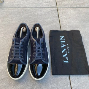 Lanvin sneakers i mörkblå mocka - Snygga sneakers från Lanvin i mörkblå mocka med lackad svart tå och vita sulor. Skorna har klassisk snörning och en stilren, rund tå. Perfekta för dig som vill ha en lyxig och unik touch på din outfit. Dustbag från Lanvin medföljer.