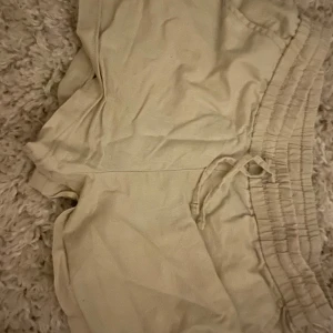 Beiga shorts från Gina Tricot - Snygga beiga shorts från Gina Tricot med elastisk midja och snörning. De är tillverkade i ett mjukt material som känns skönt mot huden och har en avslappnad passform. Perfekta för varma dagar när du vill ha något enkelt och stilrent.