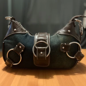 Jeans axelväska med skinn och ringdetaljer - Cool axelväska i mörkblått jeansmaterial med bruna skinnpartier och stora silvriga ringar som detaljer. Väskan har bred axelrem och kontrastsömmar, perfekt för dig som gillar en edgy och unik stil.