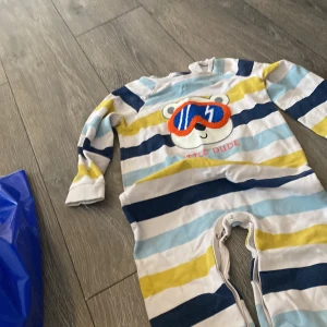 Randig body med björn från LCW baby - Supergullig långärmad body från LCW baby med färgglada ränder i vitt, blått, gult och ljusblått. Framsidan har ett broderat björnansikte med coola blå-orange glasögon och texten 'Little Dude'. Praktiska knappar i grenen för enkel påklädning.