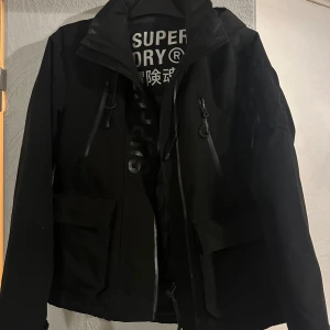 Svart vindjacka från Superdry - Svart vindjacka från Superdry med hög krage, flera dragkedjeförsedda fickor och tydlig logga på insidan. Jackan har långa ärmar och praktiska detaljer som dragsko och stora fickor framtill. Perfekt för dig som gillar streetwear och funktionell stil.