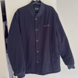 Blå collage jacka från Lyle & Scott - Svart collage jacka från Lyle & Scott Archive med röd broderad logga på bröstet och stor logga på ryggen. Jackan har ribbade muddar, knäppning med knappar och två sidofickor. Varm o go . Nypris 2000 använd 3 gånger 