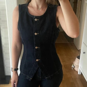 Mörkblå jeansväst från Gina Tricot - Säljer en snygg mörkblå jeansväst från Gina Tricot i storlek M. Västen har en figurnära passform med rund halsringning. Materialet är klassiskt denim med kontrastsömmar som ger en cool look. Aldrig använd.