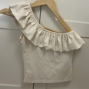 Vit oneshoulder topp med volang H&M XS - Supersnygg vit oneshoulder topp från H&M i storlek XS. Toppen har en bred volang längs halsringningen som ger en extra cool vibe. Materialet är mjukt och känns skönt mot huden. Perfekt för dig som vill sticka ut med en unik och trendig look. Skriv för mer bilder 