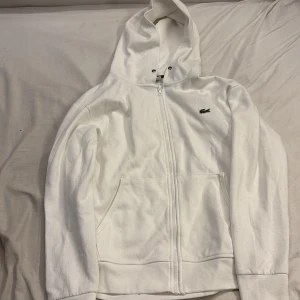 Lacoste hoodie - Vit hoodie från Lacoste i klassisk passform med dragkedja framtill och två fickor. Tröjan har huva med snörning och den ikoniska gröna krokodilloggan broderad på bröstet. Tillverkad i mjuk bomull, perfekt för en clean och stilren look. Jag kan inte garantera äktheten på denna vara