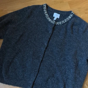 Mörkgrå kofta med pärlor från H&M - Mysig mörkgrå kofta från H&M med rund halsringning och glittrande pärldetaljer längs kragen. Koftan har knäppning framtill och är stickad i ett mjukt material. Perfekt för att lyfta din outfit med lite extra bling.