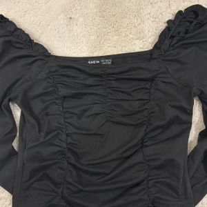 Svart långärmad topp från SHEIN - Svart långärmad topp från SHEIN i storlek S. Toppen har rynkade detaljer framtill och puffiga axlar som ger en trendig look. Modellen är figurnära med offshoulder-skärning och tillverkad i ett stretchigt syntetmaterial.