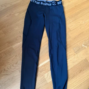 Svarta Better Bodies leggings M - Snygga svarta leggings från Better Bodies med bred resår i midjan och logga runtom. Tight passform och stretchigt material som passar perfekt till träning. Materialet är syntet och de har en sportig look.