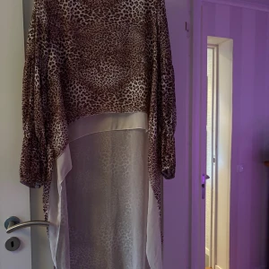 Leopardmönstrad blus med lång baksida från Chica London - Säljer en transparent blus med lång baksida från Chica London i leopardmönster. Klänningen har vida ärmar och är tillverkad i 100% polyester. Den har en snygg asymmetrisk design med ljusa inslag och passar perfekt för dig som vill sticka ut.