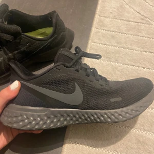 Svarta Nike React sneakers - Svarta Nike React sneakers med grå sula och stort grått swoosh på sidan. Ovandelen är i mesh för bra andningsförmåga och skon har snörning. 