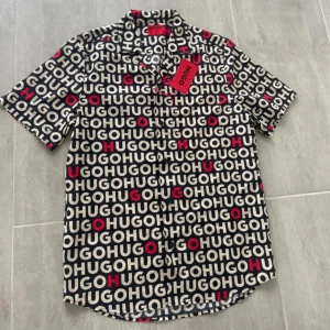 Svart kortärmad skjorta från Hugo - Snygg skjorta från Hugo med svart bas och stort logomönster i vitt och rött över hela plagget. Skjortan har klassisk krage, knäppning framtill och korta ärmar. Perfekt för dig som vill sticka ut med en trendig look. Prislapp siter kvar 