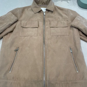 Beige mocka overshirt med dragkedja - Stilren beige overshirt från Bershka i mjuk mocka-look. Jackan har två bröstfickor med lock, snedställda fickor med dragkedja och hel dragkedja framtill. Perfekt för lager-på-lager och snygg till både jeans och chinos.