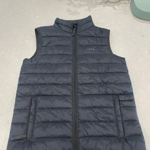 Svart dunväst från OCN Wear - Säljer en svart dunväst från OCN Wear med quiltad design och ståkrage. Västen har dragkedja framtill och två fickor med knappstängning. Perfekt för lager-på-lager och chill stil. Passar dig som gillar enkel och funktionell look. Barn-storlek 134-140.