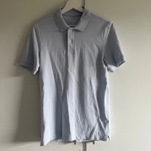 Ljusblå pikétröja från Dressmann - Säljer en stilren ljusblå pikétröja från Dressmann i storlek S. Tröjan har klassisk krage, knäppning med tre knappar och är kortärmad. Tillverkad i mjuk bomull som känns skön mot huden. Perfekt för en clean och enkel look.