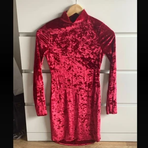 Röd sammetsklänning med polokrage - Säljer en röd sammetsklänning med polokrage och lång ärm. Klänningen är figurnära och har en kort längd, perfekt för dig som vill sticka ut med en glansig och lyxig känsla. Materialet är mjukt och skimrande sammet som fångar ljuset snyggt.