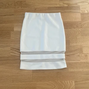 Vit kortkjol med meshdetaljer - Snygg vit kortkjol med raka linjer och två genomskinliga meshpaneler längst ner. Kjolen har en tight passform och är perfekt för dig som gillar en stilren och modern look. Materialet känns mjukt och stretchigt, vilket gör den bekväm att bära hela dagen.