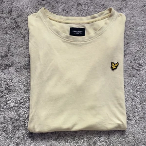 Beige t-shirt från Lyle & Scott - Snygg beige t-shirt från Lyle & Scott med klassisk rund hals och ikoniska broderade gula örnen på bröstet. Tillverkad i mjuk bomull, perfekt för en clean och stilren look.