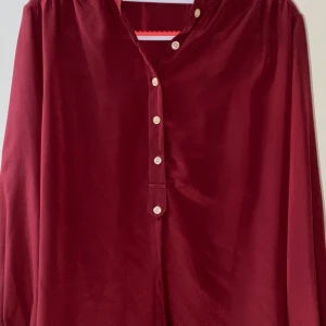 Vinröd henley-skjorta i satinlook - Snygg vinröd henley-skjorta med lång ärm och glansig satinlook. Skjortan har klassiska knappar framtill och en avslappnad passform. Perfekt för dig som vill sticka ut med en djup färg och mjukt material.