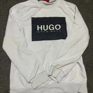 Vit sweatshirt från Hugo Boss - Vit sweatshirt från Hugo Boss med svart rektangulär logga på bröstet. Tröjan har rund halsringning, långa ärmar och ribbade muddar. Klassisk och stilren design som passar perfekt till jeans eller joggers.