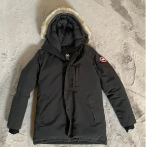 Canada Goose chateau  - Säljer en svart dunparkas från Canada Goose med huva och snygg pälsdetalj. Jackan har flera fickor, dragkedja och knappar framtill samt klassisk logopatch på ärmen. Perfekt för kalla dagar och riktigt stilren look.