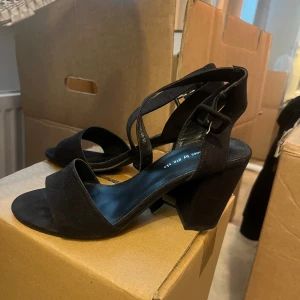 Svarta sandaler med klack - Svarta sandaler med bred rem över tårna och justerbar rem runt vristen. Skorna har öppen tå och en stabil, hög klack. Perfekta för dig som gillar stilrena och klassiska skor.