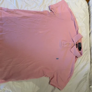 Polo Ralph Lauren  - Säljer en klassisk rosa pikétröja från Polo Ralph Lauren i storlek L. Tröjan har korta ärmar, krage med knappar och den ikoniska blå broderade loggan på bröstet. Modellen är custom fit och materialet är mjuk bomull. Hör av dig vid fler frågor 