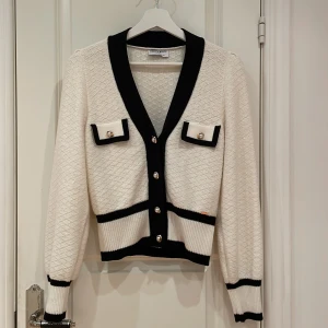 Vit kofta från Holly & Whyte med svarta detaljer - Snygg vit kofta från Holly & Whyte med svarta kontrastkanter och pärlknappar. Koftan har två dekorativa fickor framtill, ribbade muddar och v-ringad hals. Perfekt för lager-på-lager och ger en chic vibe till din outfit.