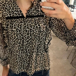 Leopardmönstrad blus med volang - Säljer en snygg blus med leopardmönster i beige och svart. Blusen har volangdetaljer längs axlarna och svarta band framtill. Långärmad modell med lätt transparent tyg och avslappnad passform.