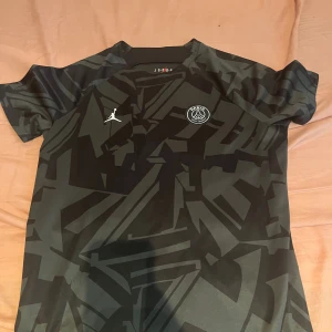 PSG x Jordan svart fotbollströja L - Snygg svart PSG fotbollströja från Jordan med grått grafiskt mönster. Tröjan har korta ärmar, rund hals och är tillverkad i ett lätt och ventilerande material. PSG-logga och Jumpman-logga på bröstet. Perfekt för dig som gillar streetstyle och fotboll.