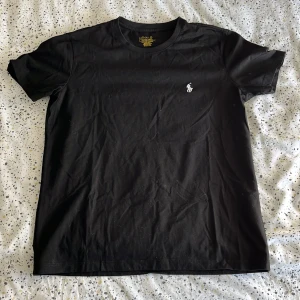Svart t-shirt från Polo Ralph Lauren - Svart t-shirt från Polo Ralph Lauren med klassisk vit broderad logga på bröstet. Modellen är slim fit och har rund halsringning och korta ärmar. Tillverkad i mjuk bomull för en skön känsla. Perfekt till jeans eller shorts för en clean och stilren look.