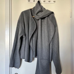 Grå cardigan/jacka från Gina Tricot - Säljer en gråmelerad cardigan/jacka från Gina Tricot i storlek S. Jackan har en lös passform, lång ärm och medföljande scarf.  Mjukt material, snygga kantdetaljer. Ingen knäppning framtill. Oanvänd, prislappen kvar. 