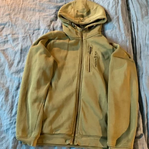 Olivgrön hoodie från Sail Racing - Olivgrön hoodie från Sail Racing med dragkedja hela vägen, huva med snörning och en bröstficka med dragkedja. Broderad logga på huvan och bröstet. Mjuk insida och långa ärmar, perfekt för kyliga dagar.