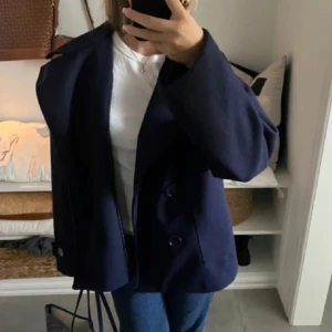 Mörkblå kavaj med knappar - Snygg mörkblå kavaj med klassisk passform och stora knappar framtill. Jackan har lång ärm och är tillverkad i ett mjukt ullmaterial som ger en stilren look. Perfekt att slänga över en t-shirt för en chill men uppklädd vibe.