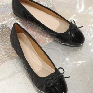 Svarta quiltade ballerinaskor med rosett - Snygga svarta ballerinaskor med quiltad design och lackad tå. Skorna har en söt rosett framtill och rund tå. Perfekta för dig som gillar klassisk och stilren look. Tillverkade i skinnliknande material.