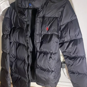 Svart pufferjacka från Polo Ralph Lauren - Svart pufferjacka från Polo Ralph Lauren med röd broderad logga på bröstet. Jackan har hög krage, dragkedja framtill och två sidofickor med dragkedja. Perfekt för kalla dagar och riktigt snygg till streetwear-stilen. Passar bra, snart vinter 🥶 