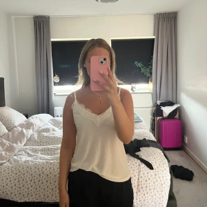 Vit linnetopp med spetsdetalj - Säljer en vit linnetopp med tunna axelband och v-ringning dekorerad med spets. Toppen har en mjuk och flowy passform, perfekt för varma dagar. Enkel och stilren design som är lätt att matcha.