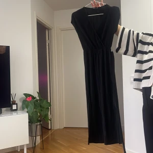 Svart omlottklänning i lång modell - Snygg svart långklänning med omlottliv och v-ringning. Klänningen har en markerad midja och är ärmlös, vilket ger en elegant och stilren look. Perfekt för dig som gillar klassiska och tidlösa plagg.