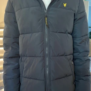 Mörkblå pufferjacka från Lyle & Scott - Säljer en mörkblå pufferjacka från Lyle & Scott med huva och dragkedja framtill. Jackan har en gul logga på bröstet och två fickor på sidorna. Perfekt för kalla dagar och riktigt skön att bära.