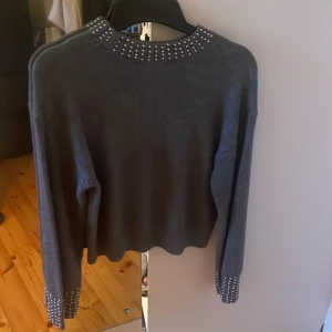 Mörkgrå stickad tröja med pärldetaljer - Mörkgrå stickad tröja med långa ärmar och rund halsringning. Snygga pärldetaljer runt halsen och vid ärmsluten ger en extra touch. 