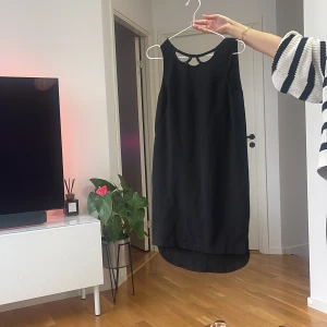 Svart ärmlös klänning med pärldetaljer - Svart ärmlös klänning med snygg v-ringning framtill dekorerad med svarta pärlor. Klänningen har en längre bakdel och en cool cut-out detalj i ryggen. Perfekt för dig som gillar stilrena och eleganta plagg med en twist.