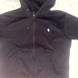 Svart hoodie från Ralph Lauren - Svart hoodie från Ralph Lauren med klassisk vit broderad logga på bröstet. Tröjan har dragkedja framtill, huva med snörning och två fickor. Perfekt för en chill och stilren look.